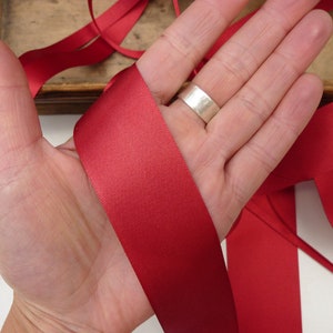 Puede incluir: Primer plano de una mano sosteniendo una cinta de raso roja. La cinta tiene una textura suave y brillante. Otras longitudes de cinta son visibles en el fondo. La imagen sugiere un tema de artesan&iacute;a o envoltura de regalos.