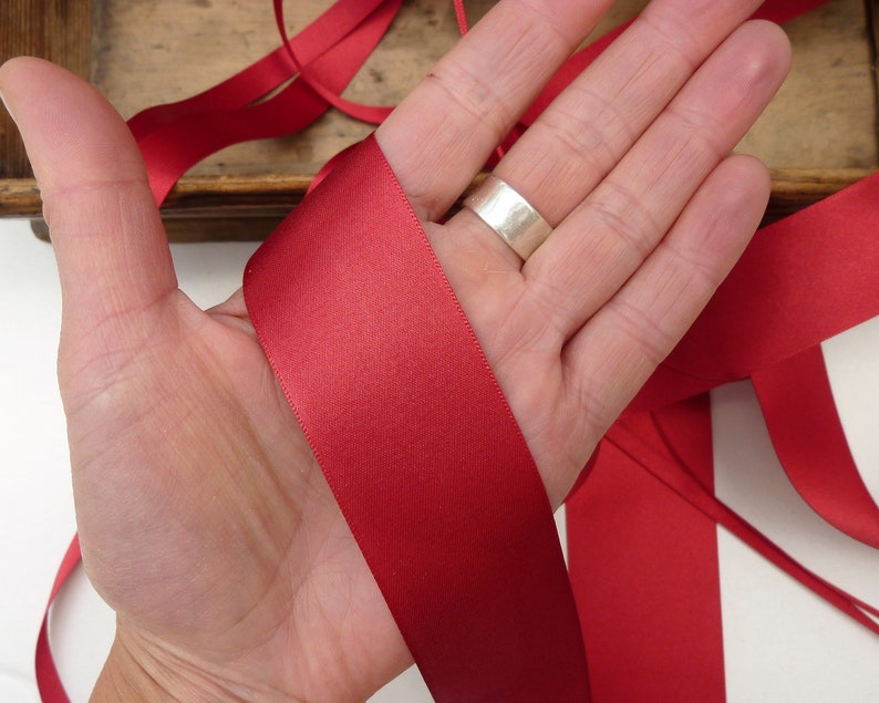 Puede incluir: Primer plano de una mano sosteniendo una cinta de raso roja. La cinta tiene una textura suave y brillante. Otras longitudes de cinta son visibles en el fondo. La imagen sugiere un tema de artesan&iacute;a o envoltura de regalos.