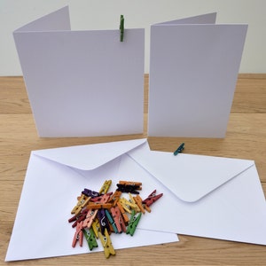 May include: Blank white greeting cards and envelopes, held by small, colourful wooden pegs. A collection of multi-coloured mini pegs are scattered near the envelopes. The cards are open, ready for a message.