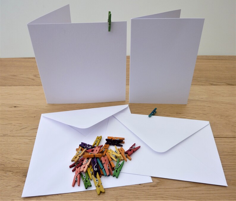 May include: Blank white greeting cards and envelopes, held by small, colourful wooden pegs. A collection of multi-coloured mini pegs are scattered near the envelopes. The cards are open, ready for a message.