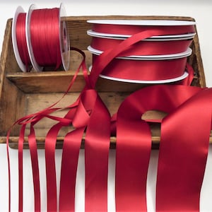 Puede incluir: Carretes de cinta de raso roja y cinta suelta en una bandeja de madera r&uacute;stica. Los carretes son blancos con cinta roja enrollada. La cinta suelta est&aacute; extendida, mostrando el brillo del material.
