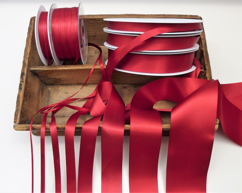Puede incluir: Carretes de cinta de raso roja y cinta suelta en una bandeja de madera r&uacute;stica. Los carretes son blancos con cinta roja enrollada. La cinta suelta est&aacute; extendida, mostrando el brillo del material.