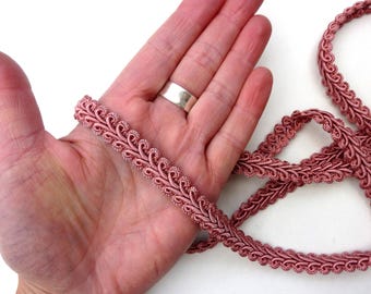 Könnte beinhalten: Nahaufnahme einer Länge rosafarbenen Zierborte mit einem dekorativen, geschlungenen Design. Die Borte wird in einer Hand gehalten, die die komplizierten Details und die Textur zeigt. Die Borte wird wahrscheinlich zum Nähen, Basteln oder Verschönern verschiedener Projekte verwendet.