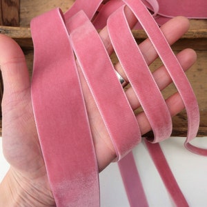Puede incluir: Cinta de terciopelo rosa, de aproximadamente 2,5 cm de ancho, en un tono rosa polvoriento suave. La cinta se muestra doblada en una mano, destacando su textura suave y su tacto lujoso.