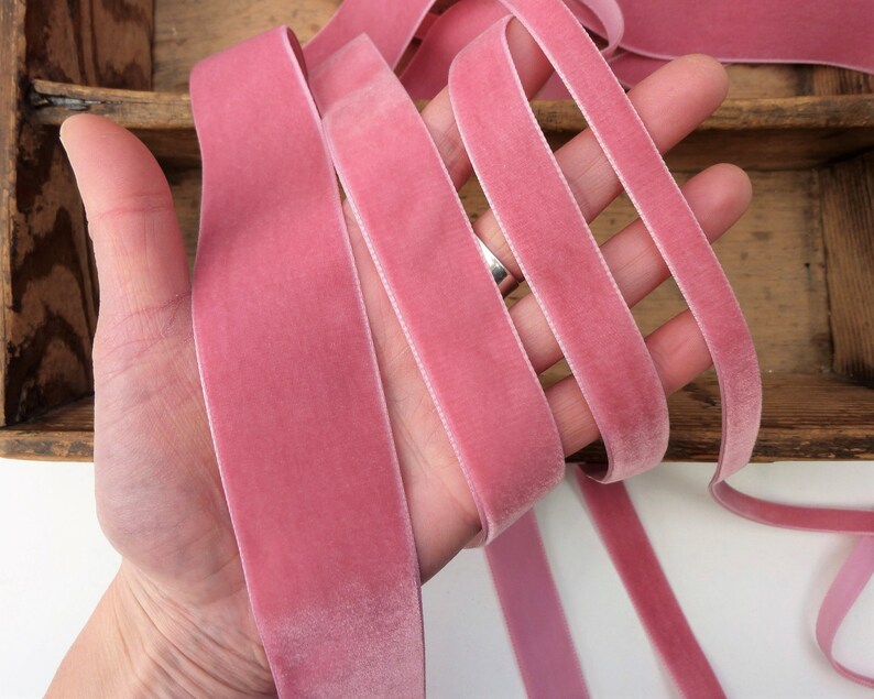 Puede incluir: Cinta de terciopelo rosa, de aproximadamente 2,5 cm de ancho, en un tono rosa polvoriento suave. La cinta se muestra doblada en una mano, destacando su textura suave y su tacto lujoso.