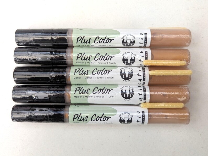 May include: Five brown and black markers with a green and white label that says "Plus Color" and "Marker | Marker | Feutres | Tusch". The label also has a black and white logo with the text "Shake Before Use" and "Kreativ".