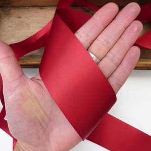 Puede incluir: Una mano sosteniendo un trozo de cinta de raso roja. La cinta es de un color rojo intenso y rico y parece suave y ligeramente reflectante. La cinta se utiliza probablemente para manualidades, envolver regalos o con fines decorativos.