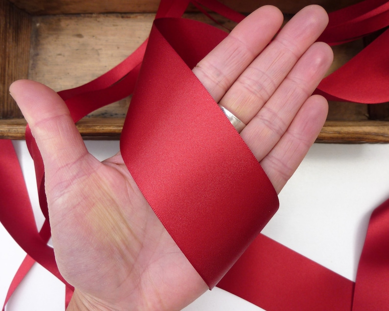 Puede incluir: Una mano sosteniendo un trozo de cinta de raso roja. La cinta es de un color rojo intenso y rico y parece suave y ligeramente reflectante. La cinta se utiliza probablemente para manualidades, envolver regalos o con fines decorativos.
