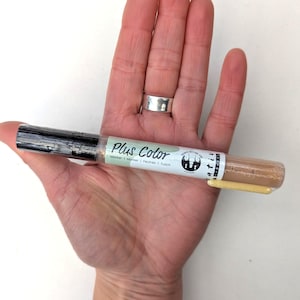 May include: A close-up of a Plus Color marker pen with a beige barrel and black cap. The pen is labeled "Plus Color Marker | Marker | Feutre | Touch" and has a green and white logo with a tree design.