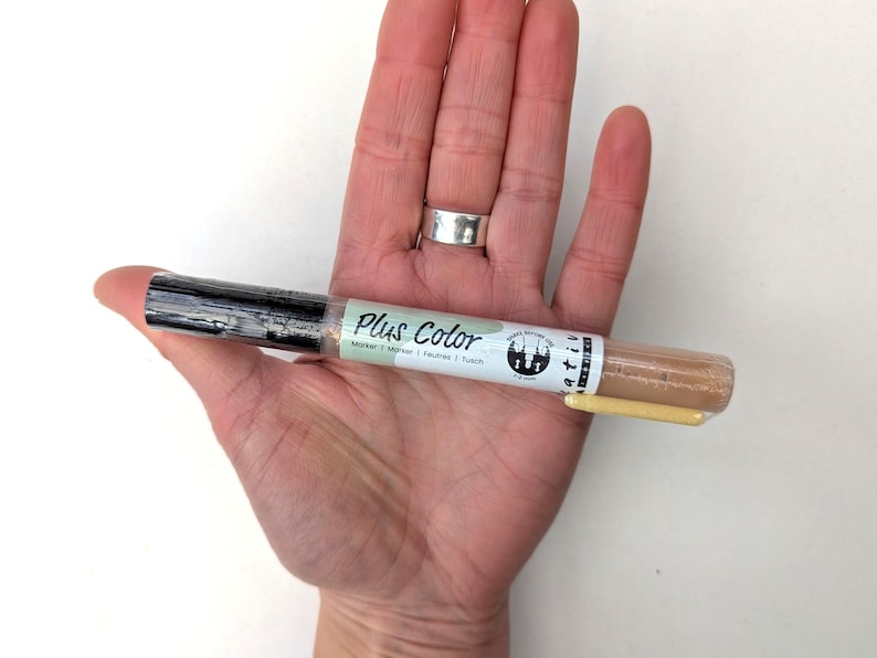 May include: A close-up of a Plus Color marker pen with a beige barrel and black cap. The pen is labeled "Plus Color Marker | Marker | Feutre | Touch" and has a green and white logo with a tree design.