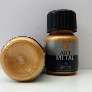K&ouml;nnte beinhalten: Zwei Flaschen goldener Metallic-Farbe. Die Flasche rechts hat einen schwarzen Deckel und ein Etikett mit der Aufschrift "Art Metal" und darunter dem Markennamen "Schjerning". Die Flasche links steht auf dem Kopf und zeigt die goldene Farbe.