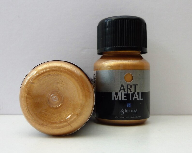 K&ouml;nnte beinhalten: Zwei Flaschen goldener Metallic-Farbe. Die Flasche rechts hat einen schwarzen Deckel und ein Etikett mit der Aufschrift "Art Metal" und darunter dem Markennamen "Schjerning". Die Flasche links steht auf dem Kopf und zeigt die goldene Farbe.