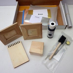 K&ouml;nnte beinhalten: Ein Bastelset f&uuml;r Metallblatt-Projekte. Das Set enth&auml;lt zwei Holzbilderrahmen, eine Holzkiste, einen Holzblock, eine Flasche schwarzer Farbe, eine kleine Tasche mit einem Pinsel, einen Stift und ein Metallblatt. Auf dem Metallblatt sind die W&ouml;rter "Brass" und "Pewter" aufgedruckt.