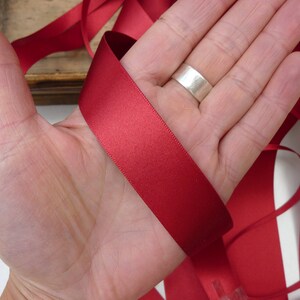Puede incluir: Un trozo de cinta de raso rojo intenso, sostenido en una mano. La cinta es lisa y tiene un ligero brillo, lo que sugiere un material de alta calidad. La cinta es adecuada para manualidades, envolver regalos o fines decorativos.
