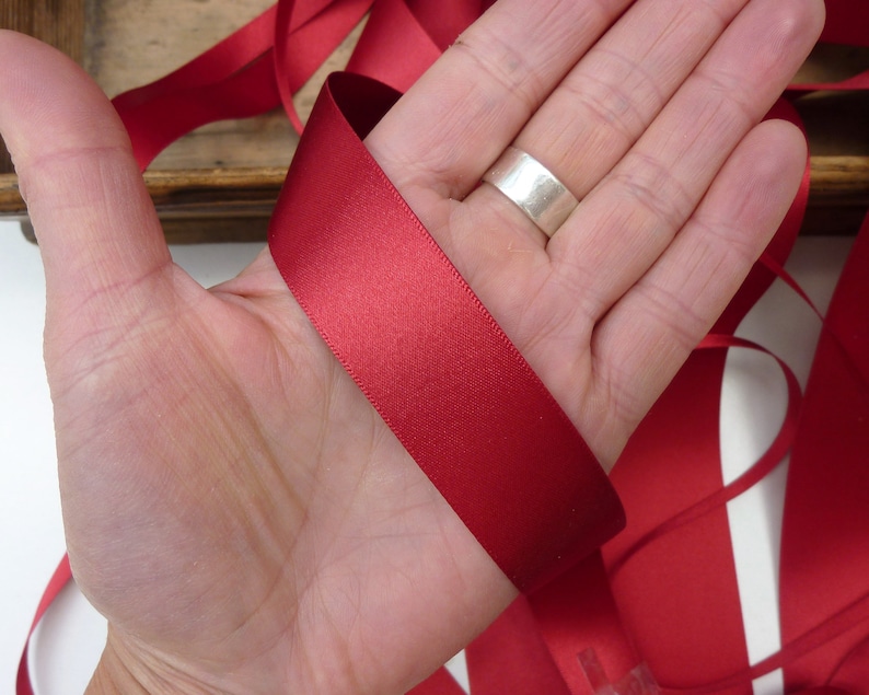 Puede incluir: Un trozo de cinta de raso rojo intenso, sostenido en una mano. La cinta es lisa y tiene un ligero brillo, lo que sugiere un material de alta calidad. La cinta es adecuada para manualidades, envolver regalos o fines decorativos.