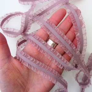 May include: A close-up of a dusty rose coloured ruffled elastic trim. The trim is soft and delicate, with a subtle sheen. It is perfect for adding a touch of elegance to any project.