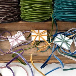 May include: Three large spools of velvet ribbon in brown, lime green, and teal are positioned above three small white rectangular boxes. Each box is tied with a thin velvet ribbon in shades of dusty pink, mustard yellow, and sage green.