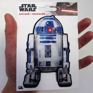 May include: A white and blue Star Wars R2-D2 iron-on patch. The patch is on a white card with the words "Star Wars" and "Appliqué / Aplicación" printed on it.