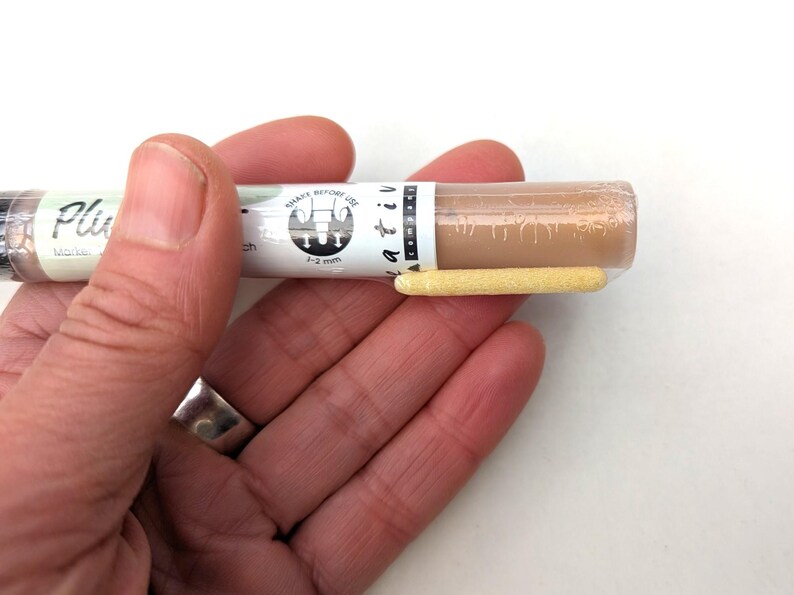 May include: A close-up of a brown paint marker with a white label that says "Shake Before Use" and "1-2 mm". The marker has a yellow tip.