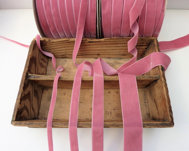 Puede incluir: Una caja de madera llena de cinta de terciopelo rosa. La cinta est&aacute; dispuesta en un patr&oacute;n decorativo y est&aacute; lista para ser utilizada para manualidades o decoraci&oacute;n.