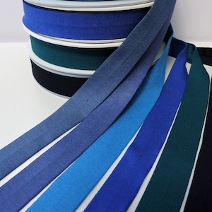 May include: A selection of bias binding in shades of blue and teal. Several spools of the tape are stacked in the background, with lengths of tape fanning out in the foreground. The colours range from light blue to navy and teal.