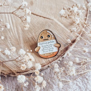 Könnte beinhalten: Eine kartoffelförmige Anstecknadel mit dem Text "Positive potato" und einer motivierenden Botschaft. Die Anstecknadel ist braun und orange mit einem weißen Schild. Die Anstecknadel ist von Trockenblumen auf einer Holzoberfläche umgeben.