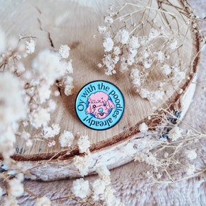 Könnte beinhalten: Runder Emaille-Pin mit hellblauem Hintergrund und einer rosa Pudel-Illustration. Der Pin trägt die Aufschrift "Oy with the poodles already!" in einem kreisförmigen Design. Der Pin ist auf einer Holzscheibe mit weißen Blumen platziert.