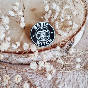Könnte beinhalten: Ein schwarz-weißer Emaille-Pin mit den Worten "BASIC WITCH" in einem kreisförmigen Design. Der Pin zeigt eine Hexe mit Hut und ist von weißen Sternen umgeben. Der Pin befindet sich auf einer Holzoberfläche mit weißen Blumen.