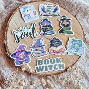 Könnte beinhalten: Eine Sammlung farbenfroher Aufkleber mit einem Hexen- und Bücher-Thema. Designs umfassen einen Geist mit Büchern, offene Bücher mit Federn, eine Hexe mit Büchern und die Wörter "bookish soul" und "book witch". Die Aufkleber sind auf einer Holzoberfläche angeordnet.