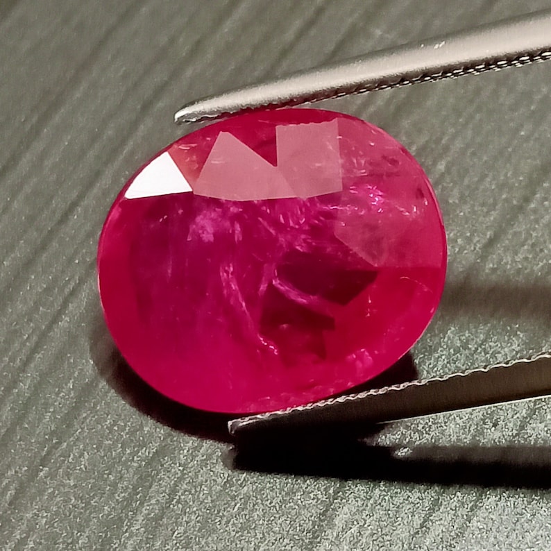 May include: A vibrant, deep red oval-shaped gemstone with a smooth, polished surface. The gemstone is held in place by a pair of tweezers.