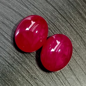 May include: Two oval-shaped, bright red gemstones. The gemstones are smooth and polished, with a high gloss finish.
