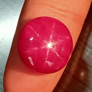 Può includere: Una gemma di rubino stellato a taglio cabochon rotondo con una vibrante tonalità rosso-rosa intenso. La pietra mostra un distinto effetto di asterismo a sei raggi, dove la luce si riflette in un motivo a stella sulla superficie liscia e bombata.