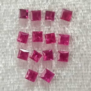 May include: A collection of 12 square-shaped, faceted, ruby red gemstones. The gemstones are arranged in three rows on a white, textured surface.