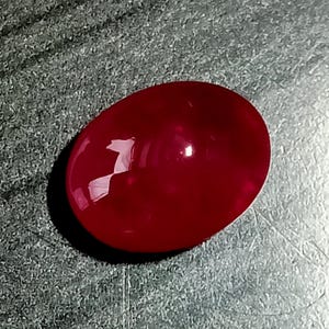 Könnte beinhalten: Ein polierter, oval geformter Edelstein in einer tiefen, gesättigten roten Farbe. Die Oberfläche des Steins ist glatt und abgerundet und weist ein hochglänzendes Finish auf, das Licht reflektiert und einen hellen weißen Glanzpunkt erzeugt.