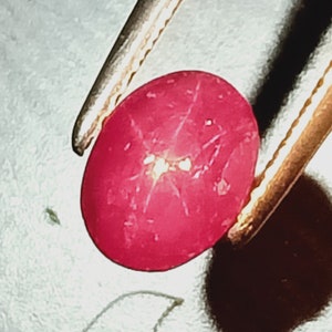 May include: A close-up of a red star ruby gemstone held by tweezers. The ruby has a star-shaped pattern in the center.