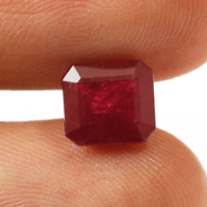 Puede incluir: Una piedra preciosa de color rojo oscuro tallada en esmeralda. La piedra preciosa es de un color rojo vibrante y tiene una superficie lisa y pulida.