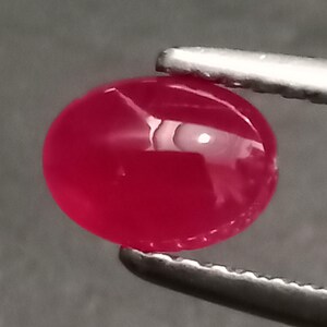 Könnte beinhalten: Ein polierter, oval geformter, tiefroter Edelstein wird von einer Pinzette vor einem grauen Hintergrund gehalten. Der Edelstein hat eine glatte, reflektierende Oberfläche, wobei sich das Licht auf seiner abgerundeten Form spiegelt. Die Pinzette ist silberfarben.