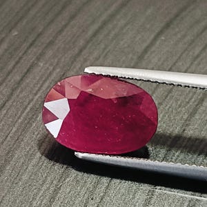以下が含まれることがあります： ピンセットで保持された、ファセット加工された楕円形のルビー。 深い赤色の宝石は、光を捉える複数のファセットを示しています。 ピンセットは銀色で、背景はテクスチャ加工された灰色の表面です。