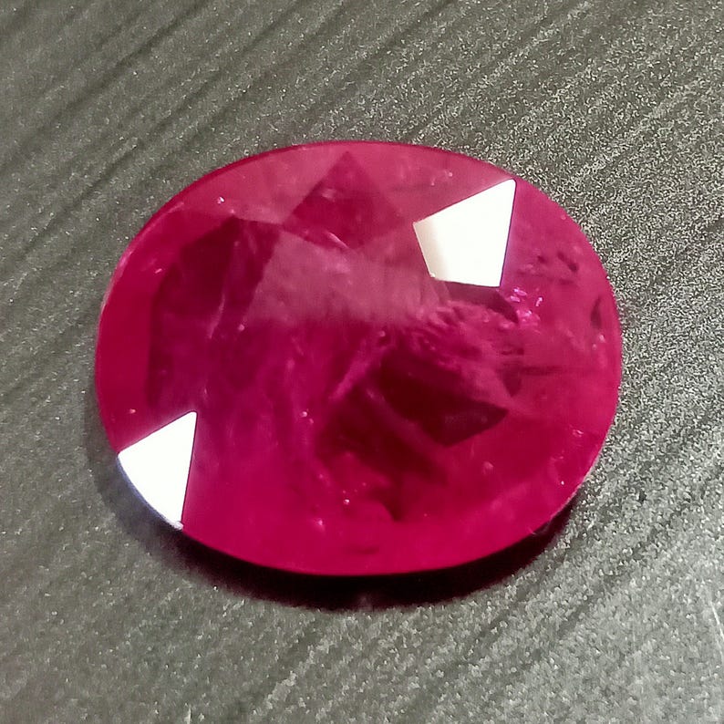 May include: A vibrant red oval gemstone with a faceted cut and white accents. The gemstone is set in a clear, polished setting.