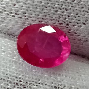 May include: A vibrant, oval-cut gemstone with a deep, saturated pinkish-red hue. The stone features multiple facets that reflect light, creating a brilliant sparkle across its surface. The gemstone is translucent, displaying internal clarity and depth.