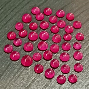 May include: A collection of 35 pink round cabochon gemstones. The gemstones are arranged in a semi-circle pattern on a dark grey surface.
