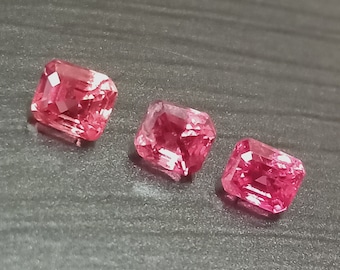 May include: Three pink gemstones cut in a square shape. The gemstones are set on a dark gray surface.