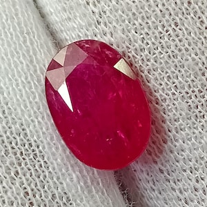 Puede incluir: Una gema de rubí de corte ovalado vibrante con un color rojo intenso y múltiples facetas que reflejan la luz. La piedra muestra una superficie pulida con inclusiones internas visibles y facetas geométricas afiladas en el borde superior.