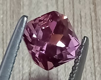 May include: A deep purple gemstone with a faceted cut. The gemstone is held in tweezers.