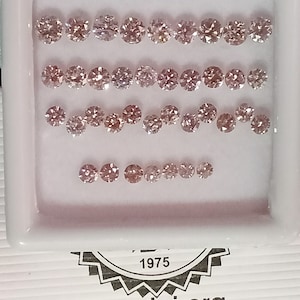 May include: A white tray with a row of 20 small, round, pink diamonds. The tray has a white background and a black border. The diamonds are arranged in four rows of five. The tray also has the text "1975" and "www.igi.org" printed on it.