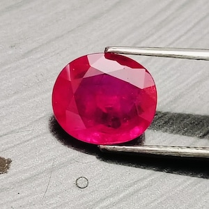 Op de afbeelding: Een levendige, ovaal geslepen robijn met een dieprode kleur en een gefacetteerd oppervlak, vastgehouden door een zilverkleurige metalen pincet. De edelsteen vertoont heldere interne structuur en scherpe geometrische facetten die licht reflecteren.
