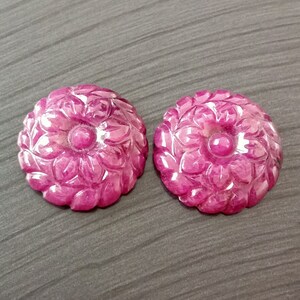 以下が含まれることがあります： 詳細な花びらの彫刻が施された、それぞれ花の形をした2つの彫刻されたルビーの宝石。宝石は濃いピンク色で、研磨された仕上げです。背景はニュートラルグレーです。