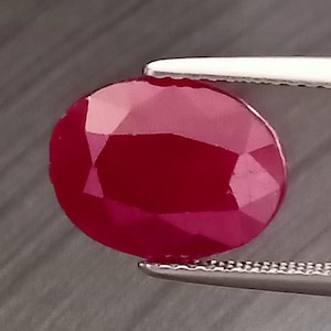 May include: An oval-cut, deep red ruby gemstone with multiple facets reflecting light. The gemstone is held by a pair of silver-toned metal tweezers, showcasing the clarity and vibrant crimson color of the stone against a dark, textured background.