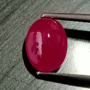 Könnte beinhalten: Ein lebhafter, tief roter ovaler Edelstein mit einer glatten, polierten Oberfläche. Der Edelstein wird von einer Pinzette gehalten.