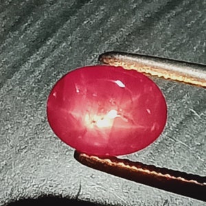 May include: A bright pink gemstone with a star-like pattern in the center. The gemstone is held by a pair of tweezers.
